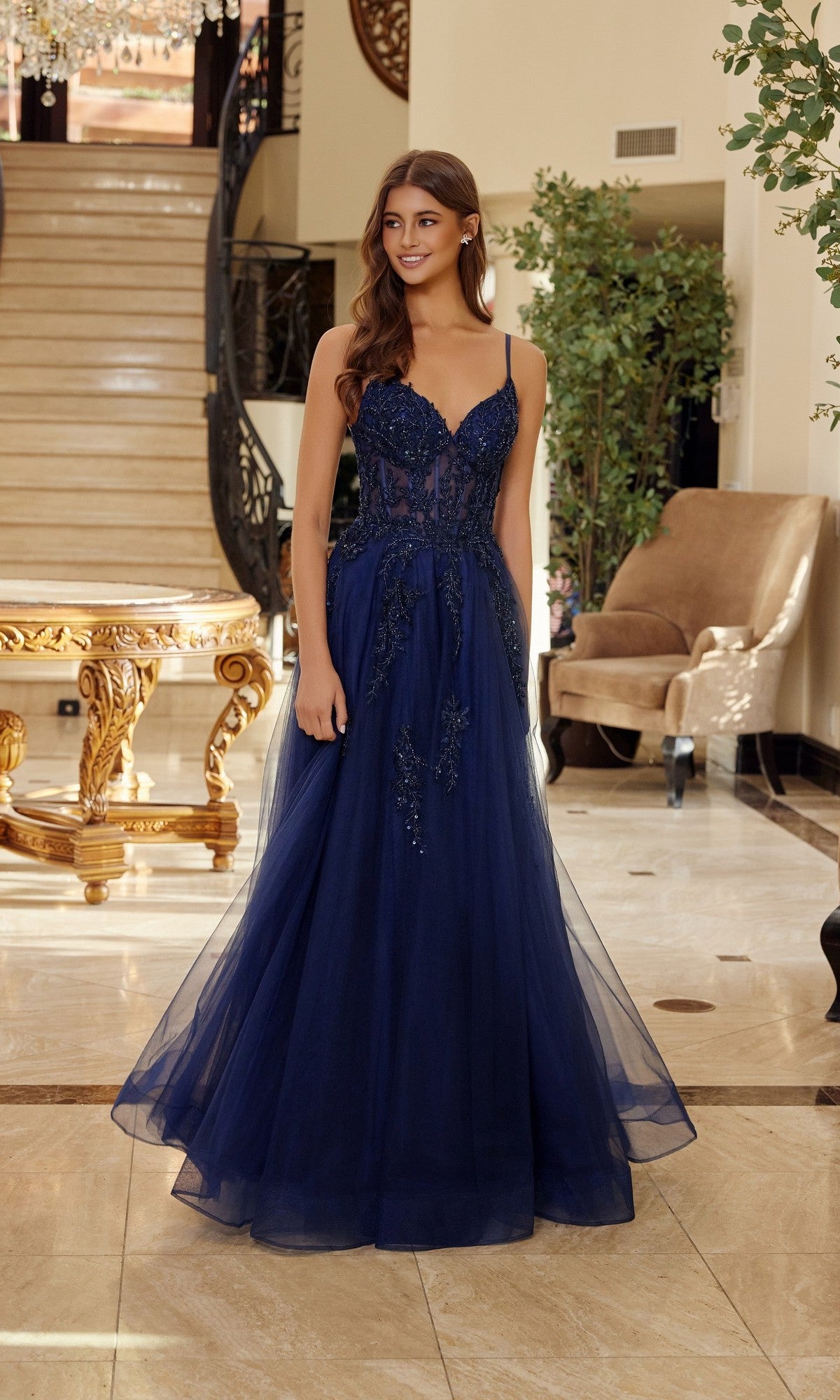 A woman stands indoors in front of a staircase and beige armchair, wearing the Nox Anabel Q1778 long prom dress in navy blue with a fitted bodice and floor-length tulle skirt.